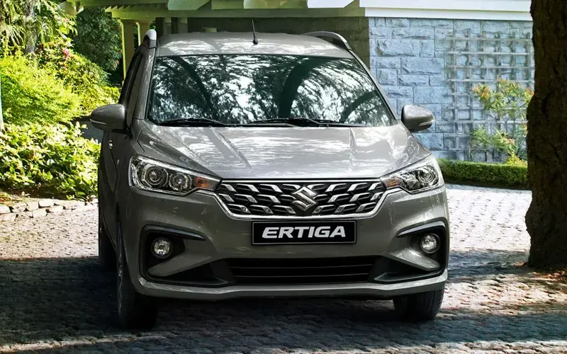 Suzuki Ertiga plateado estacionado en una entrada de vehículos, con árboles y un muro de piedra al fondo.